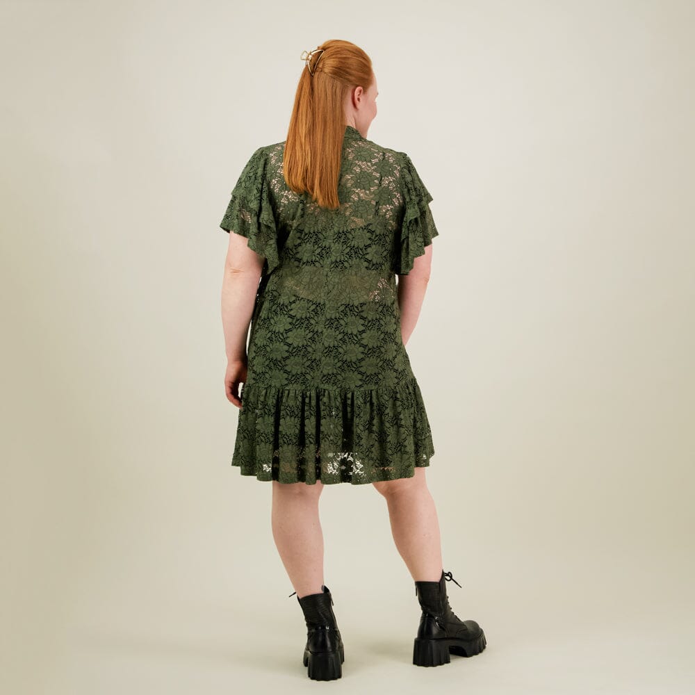 malli takaa päin jolla yllään naisten lyhyt vihreä pitsimekko frillahihoilla ja korkealla kauluksella, läpikuultava ilman aluspaitaa.