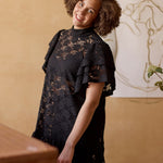 lähikuva mallista joka seisoo hieman vinottain kohti kameraa, yllään naisten lyhyt musta pitsimekko frillahihoilla ja korkealla kauluksella, läpikuultava ilman aluspaitaa.