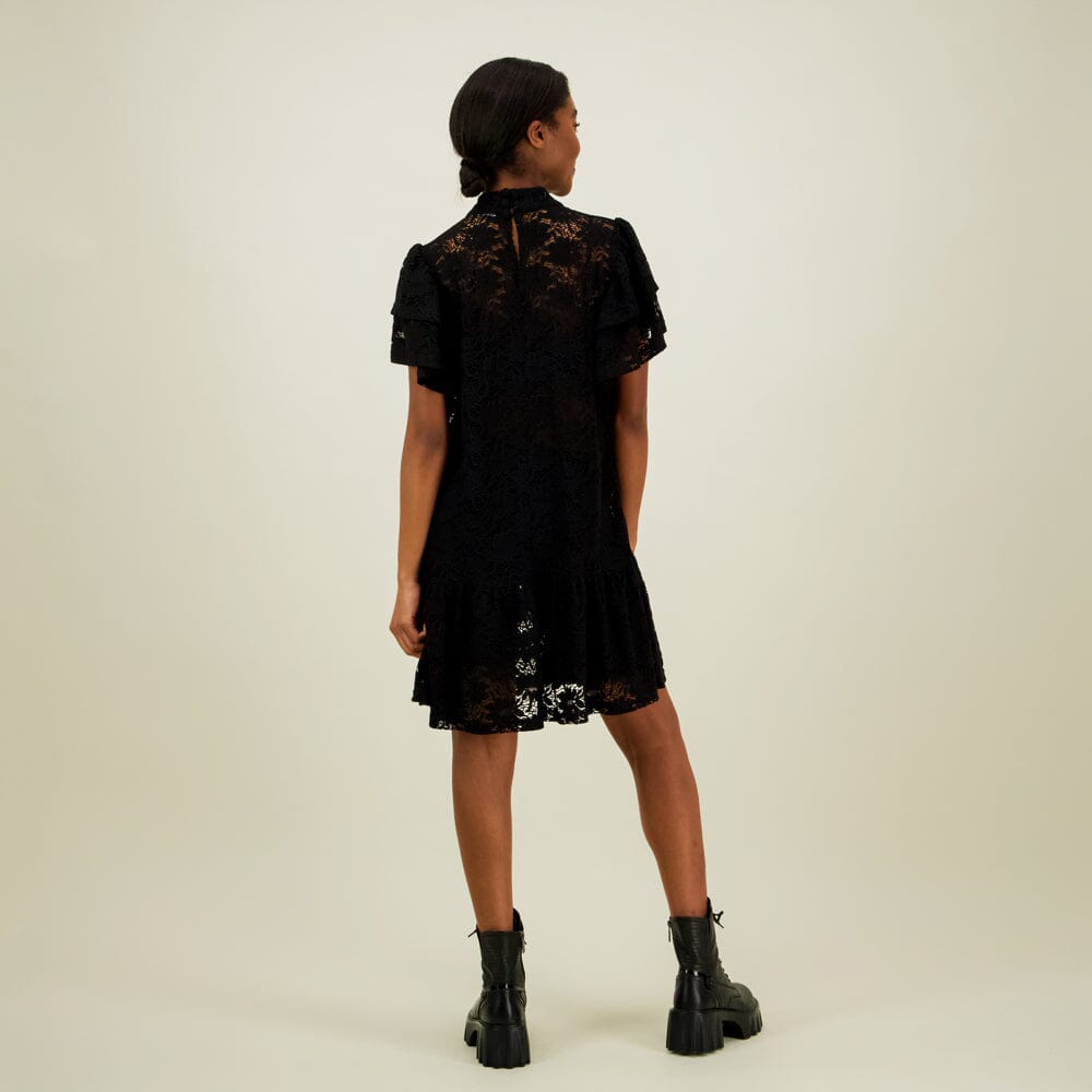 kuva mallista takaa päin jolla yllään naisten lyhyt musta pitsimekko frillahihoilla ja korkealla kauluksella, läpikuultava ilman aluspaitaa.