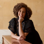 malli nojaa pianoon pitäen kättään lähellä kasvojaan, yllään naisten lyhyt musta pitsimekko frillahihoilla ja korkealla kauluksella, läpikuultava ilman aluspaitaa.