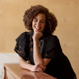 malli nojaa pianoon pitäen kättään lähellä kasvojaan, yllään naisten lyhyt musta pitsimekko frillahihoilla ja korkealla kauluksella, läpikuultava ilman aluspaitaa.