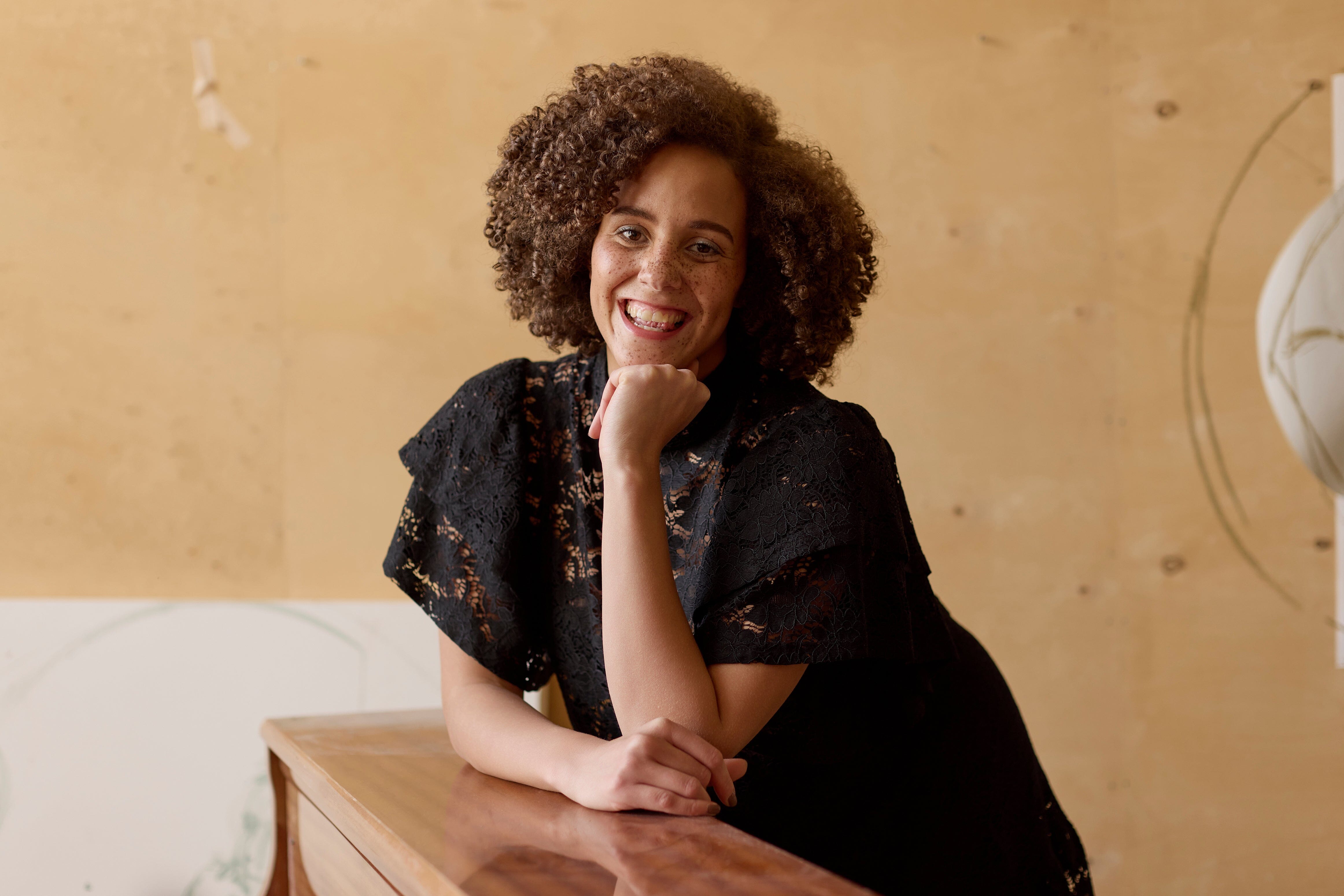 malli nojaa pianoon pitäen kättään lähellä kasvojaan, yllään naisten lyhyt musta pitsimekko frillahihoilla ja korkealla kauluksella, läpikuultava ilman aluspaitaa.