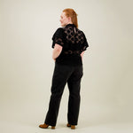 Naisten musta läpikuultava pitsitoppi korkealla kauluksella ja lyhyillä kerrostetuilla frillahihoilla, kuva mallista takaa päin. Toppi on yhdistetty mustiin farkkuihin. 