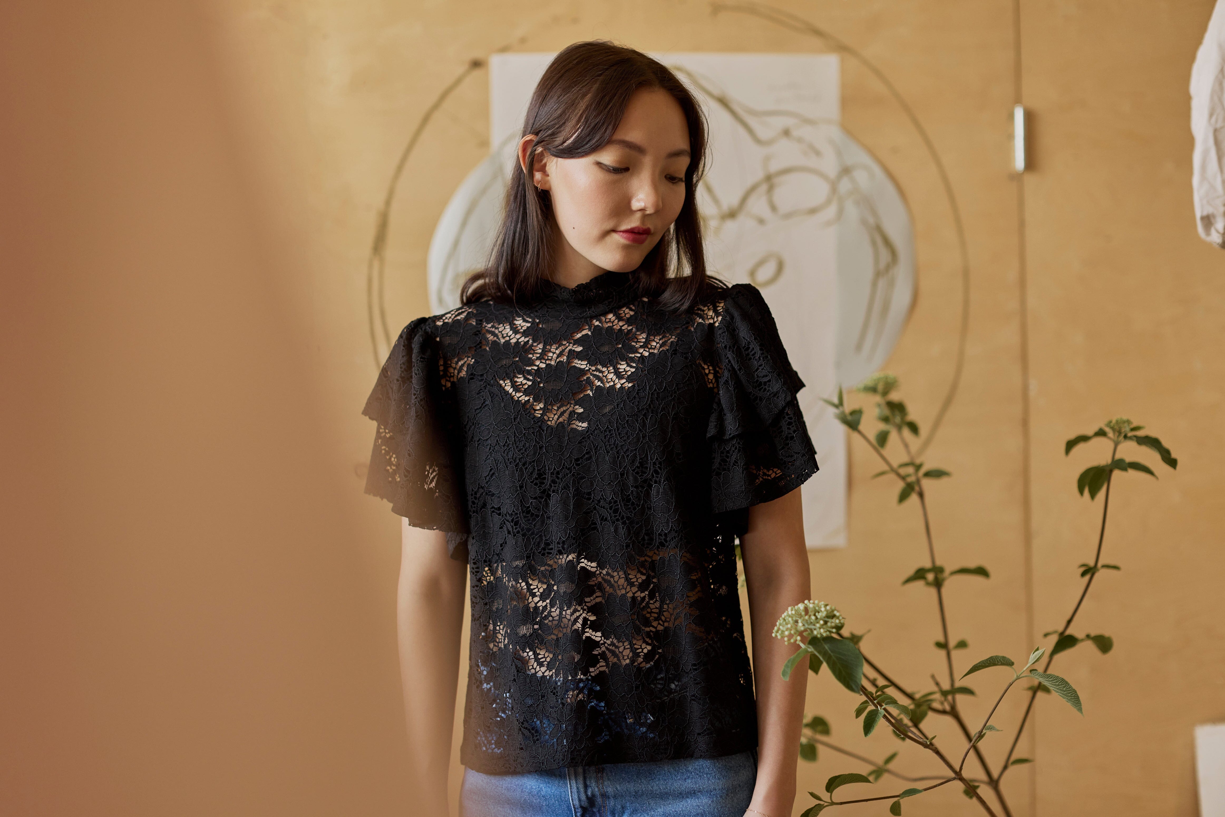 lähikuva mallista jolla yllään naisten musta läpikuultava pitsitoppi korkealla kauluksella ja lyhyillä kerrostetuilla frillahihoilla