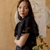 Naisten musta läpikuultava pitsitoppi korkealla kauluksella ja lyhyillä kerrostetuilla frillahihoilla, kuvassa malli lähikuvassa hieman sivuttain. 