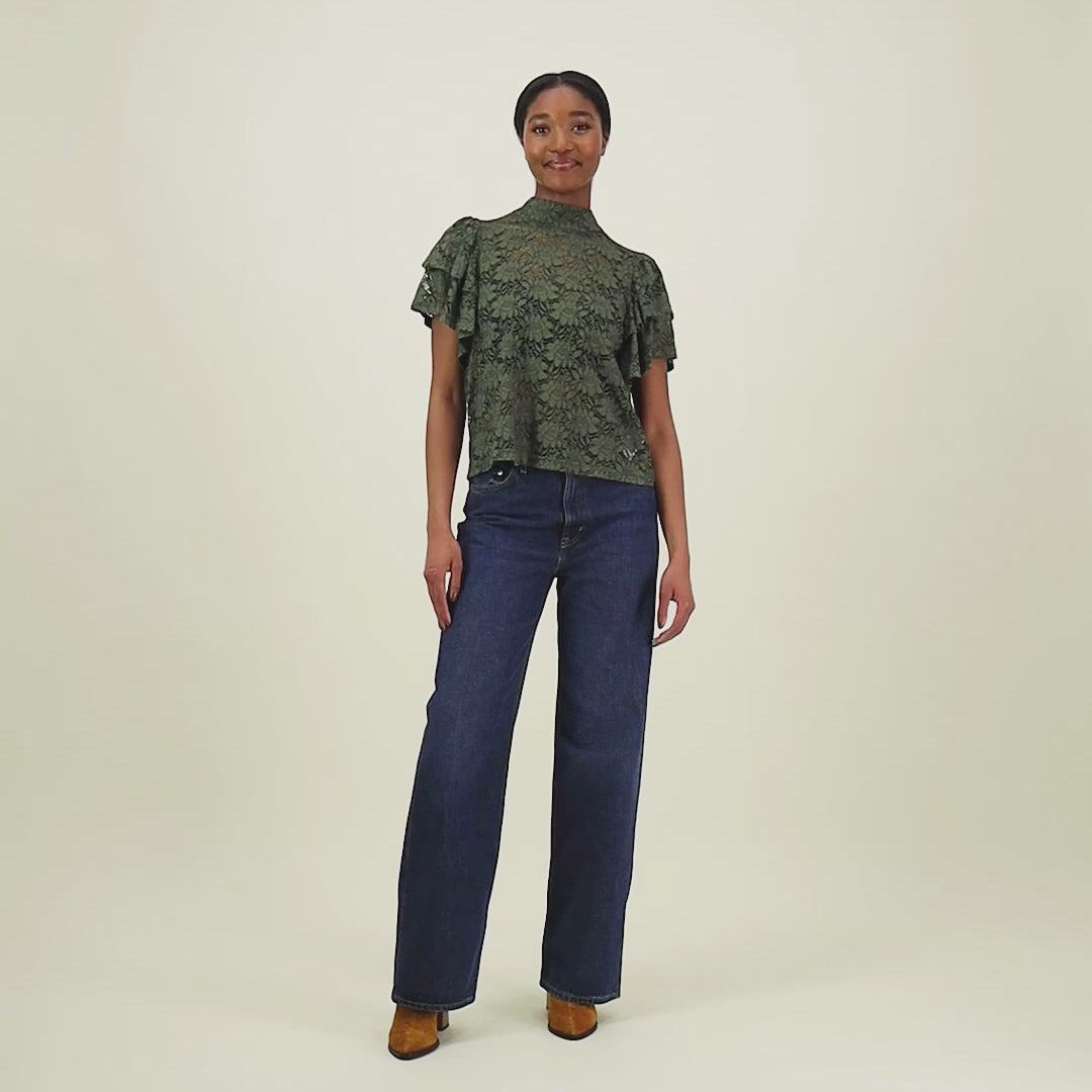 video jossa mallilla päällä vihreä läpikuultava pitsitoppi jossa korkea kaulus ja kerrostetut hihat. Toppi on yhdistetty sinisiin farkkuihin.