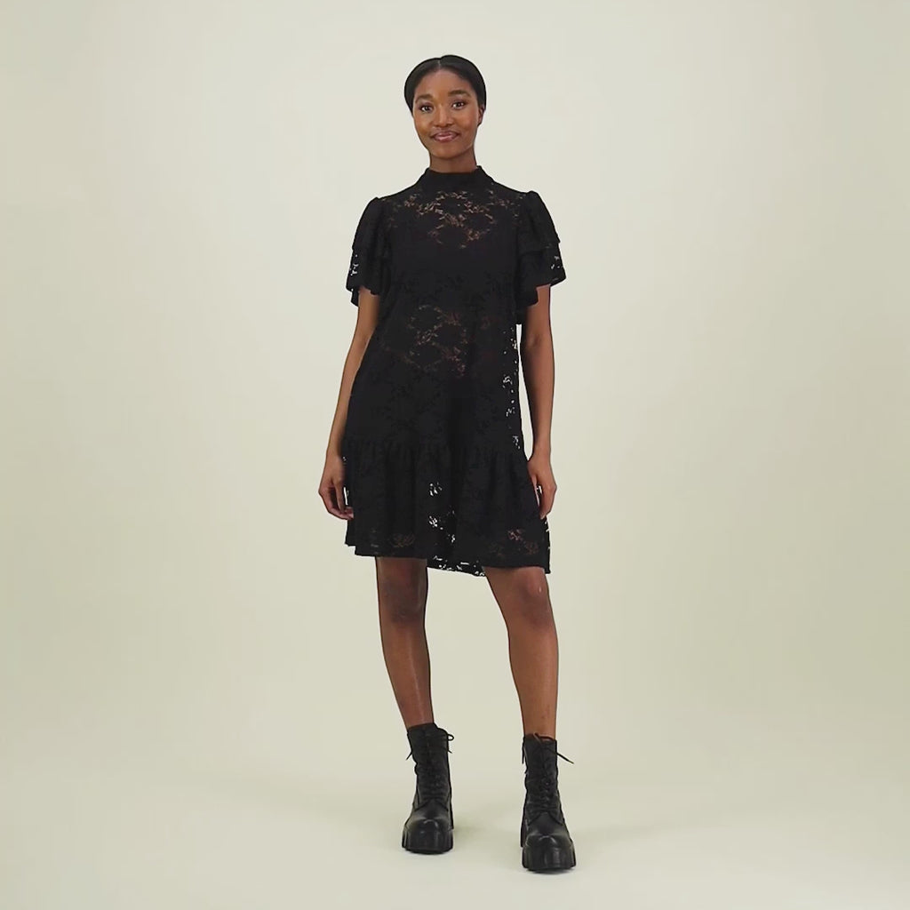 video jossa malli esittelee naisten lyhyt musta pitsimekko frillahihoilla ja korkealla kauluksella, läpikuultava ilman aluspaitaa. Mekko on yhdistetty  mustiin maihareihin. 