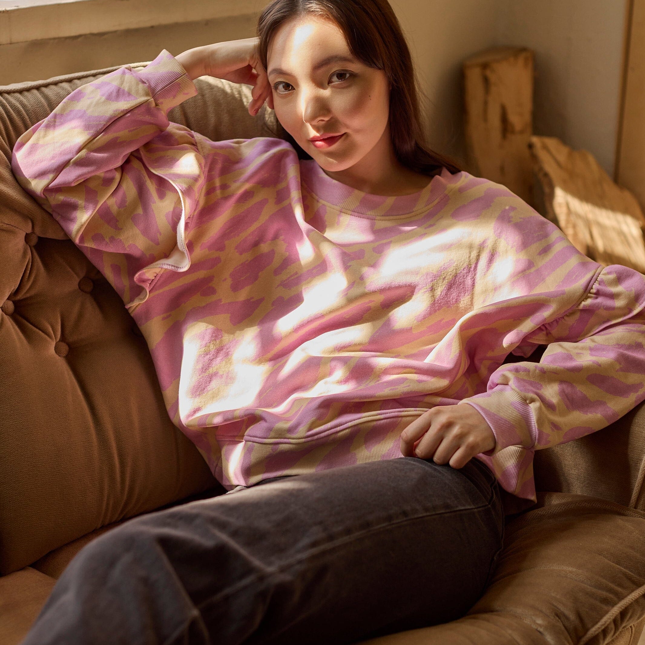 Naisten oversize-mallinen purple-sävyinen collegepaita frillahihoilla ja graafisella kuosilla, nainen istuu rennosti sohvalla, paita on yhdistetty tummiin farkkuihin. 