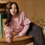 Naisten oversize-mallinen purple-sävyinen collegepaita frillahihoilla ja graafisella kuosilla, nainen istuu pöydällä. 