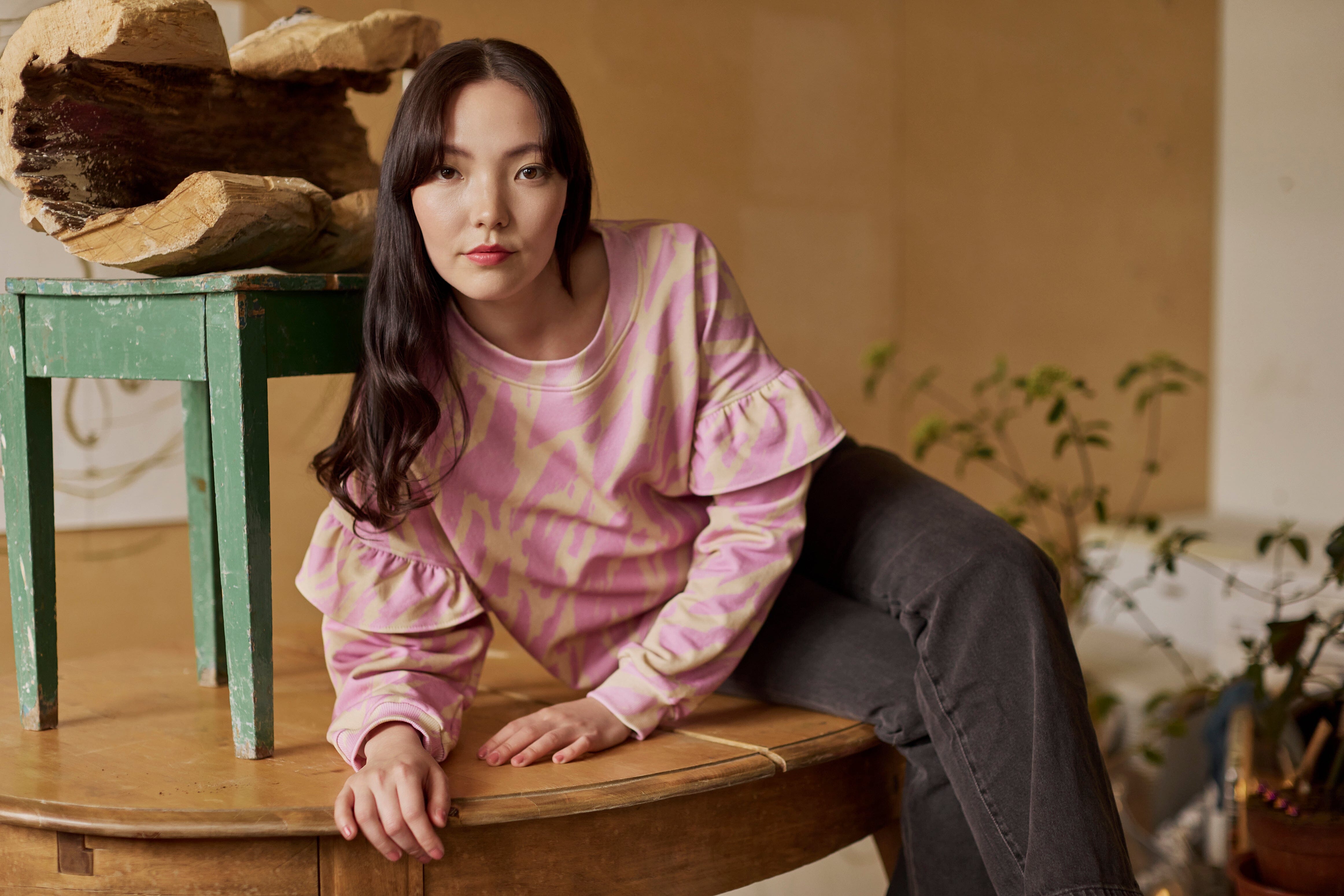 Naisten oversize-mallinen purple-sävyinen collegepaita frillahihoilla ja graafisella kuosilla, nainen istuu pöydällä. 