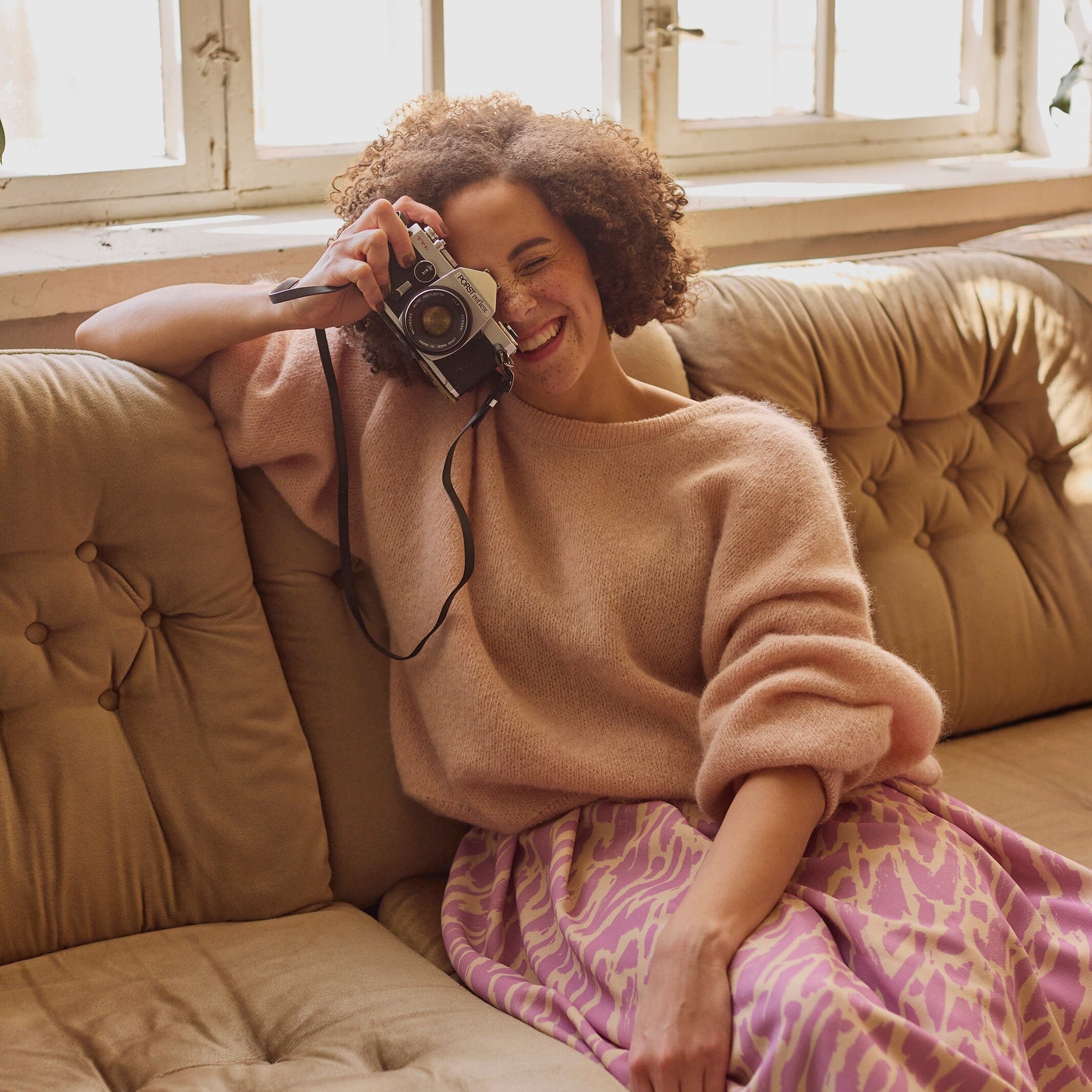 Naisten midimittainen purple-sävyinen laskostettu hame kierrätyspolyesterista, joustavalla vyötäröllä ja sivutaskuilla. Nainen istuu sohvalla katsoen kameran läpi kohti kuvaajaa.