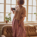 Naisten midimittainen purple-sävyinen laskostettu hame kierrätyspolyesterista, joustavalla vyötäröllä ja sivutaskuilla. Nainen seisoo sohvalla, hameeseen yhdistettynä Mohair sweater sävyssä powder. 